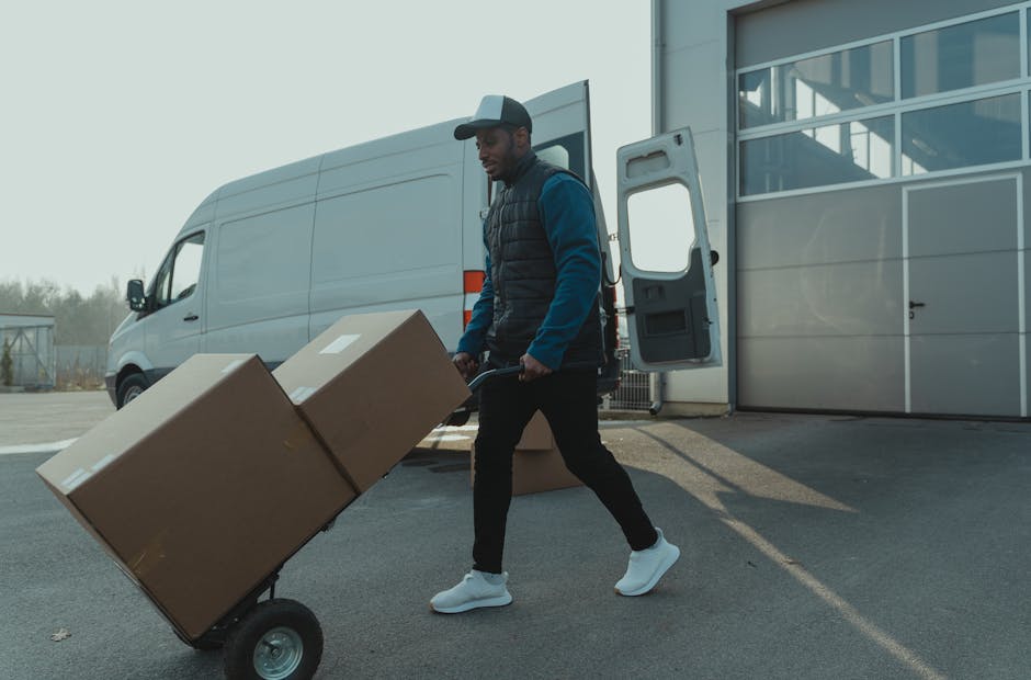 Two men, dressed in casual clothing and protective gloves, are engaged in a home relocation task outside a residential property, preparing to load a large cardboard box into the rear of a white moving