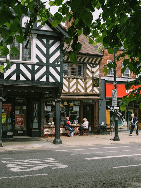 The image shows a typical street scene with two adjacent buildings featuring traditional Tudor-style black and white timber framing and brickwork, with retail shops at ground level. In front of the buildings, outdoor seating with metal tables and chairs is visible, where two elderly individuals are engaged in conversation over drinks. A woman is walking past the scene, and a man is seen walking on the pavement. A mature tree with broad green leaves partly obscures the upper portion of the buildings and provides shade. On the right side of the street, a black lamppost with a sign and a red storefront sign are visible. The street itself has painted road markings, and a parked vehicle is not visible in this segment. This scene, reflective of lively local shopping streets, aligns with the context of house removals and moving services, such as those provided by Man and a Van Bromley, emphasizing urban relocation and transport logistics.
