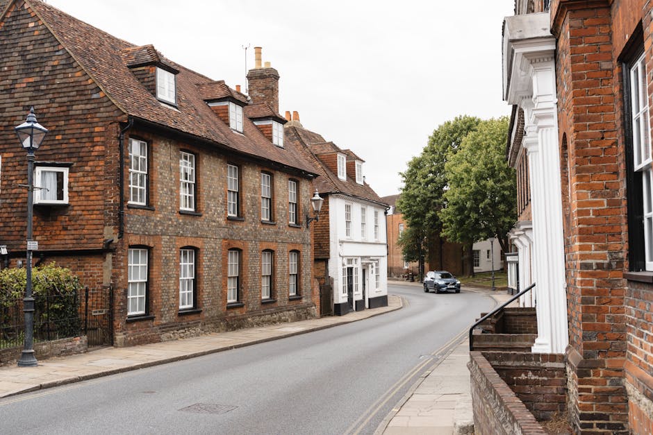 A quiet residential street scene in Bromley with traditional brick houses featuring white-framed sash windows and pitched tiled roofs. Several roadside lamp posts line the pavement, which runs alongside the houses. A modern grey car is seen driving along the narrow, gently curving road. On the right, part of a house with red brick walls and white architectural details like columns and window trims is visible. In the foreground, there are steps leading up to a doorway, and a few steps further along the pavement, there is a small section of brick wall. The background includes lush green trees and additional houses, suggesting a calm, established neighbourhood. The overcast sky results in diffuse natural lighting, typical for a grey day, suitable for visualising a typical home relocation or furniture transport process associated with [PAGE_TITLE]. [COMPANY_NAME] may use such environments for providing removals and packing services, emphasizing careful handling of household items during loading and transportation within a typical UK residential setting.